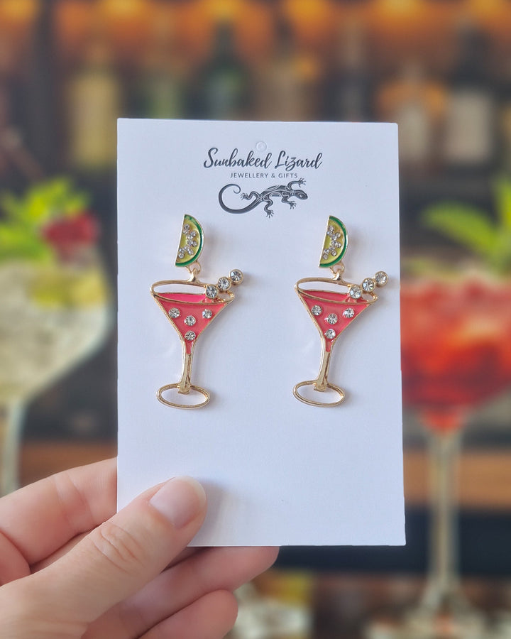 Hand holding pink cocktail earrings on a backing card against a cocktail background showing size