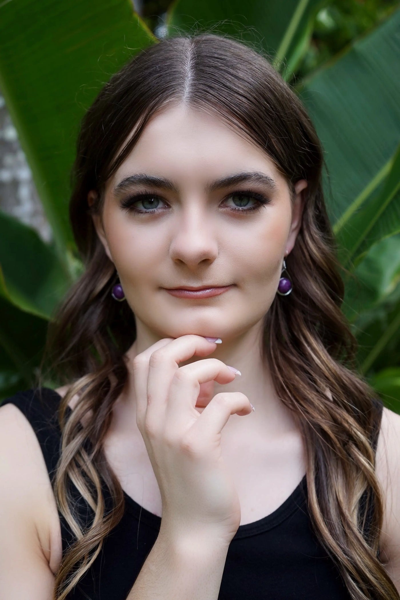 Woman with long brown hair and purple stone earrings against a green leafy background