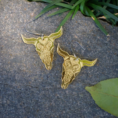 Gold bull head earrings on a stone surface with grass and leaves.