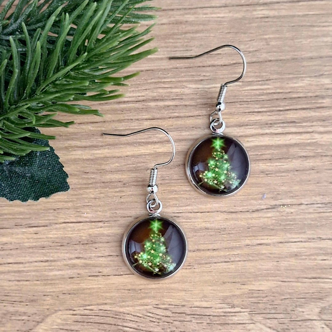 Pair of earrings with Christmas tree design on a wooden surface with greenery.