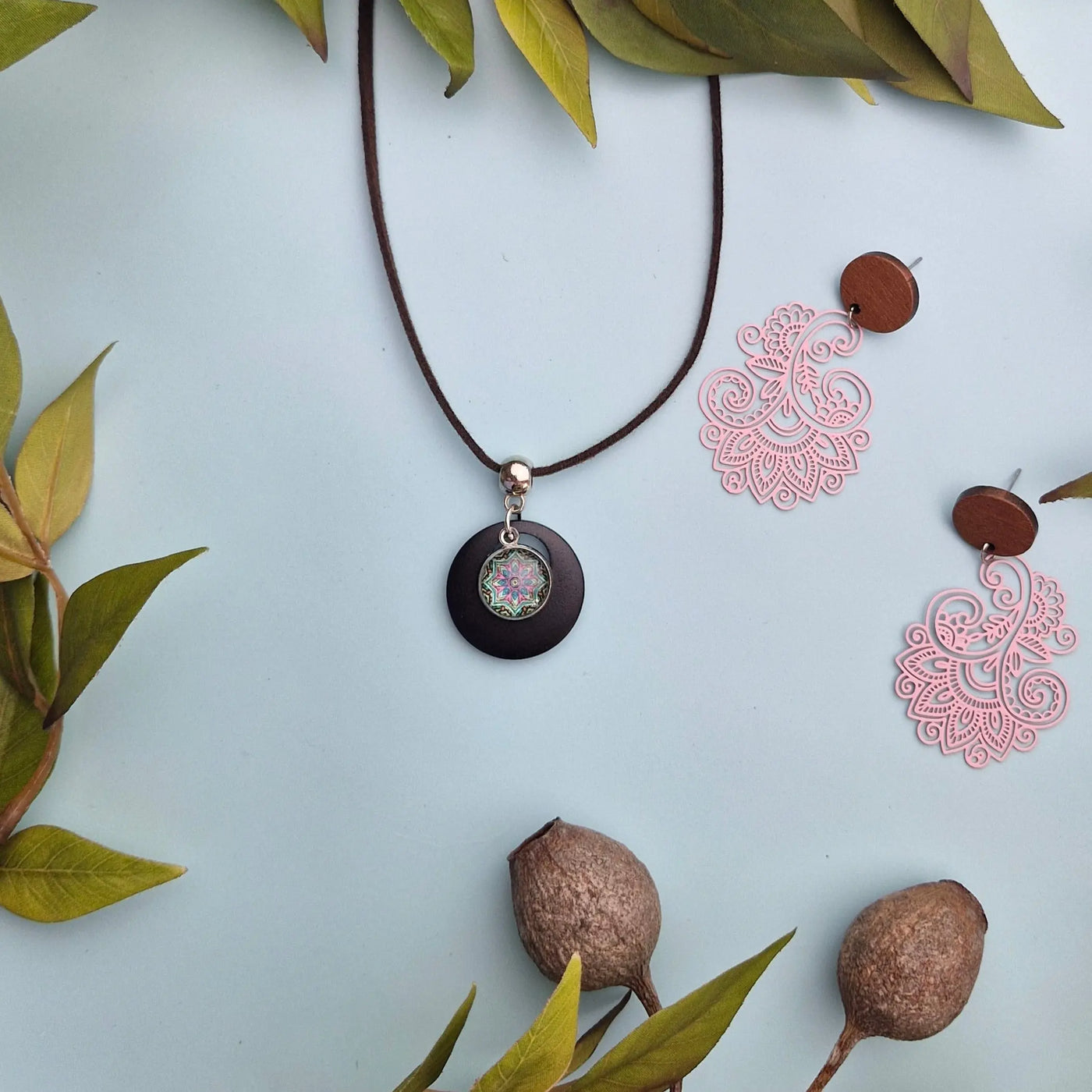 Necklace with pink floral earrings on a pale blue background surrounded by green leaves