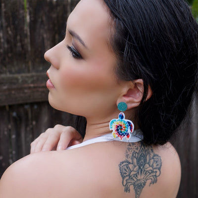 Woman wearing colorful turtle earrings with a tattoo on her shoulder against a natural background