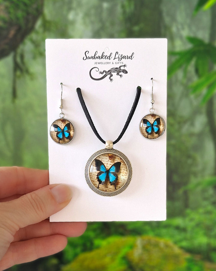 Blue Ulysses Butterfly necklace and matching drop earrings set on Sunbaked Lizard card with tropical forest background