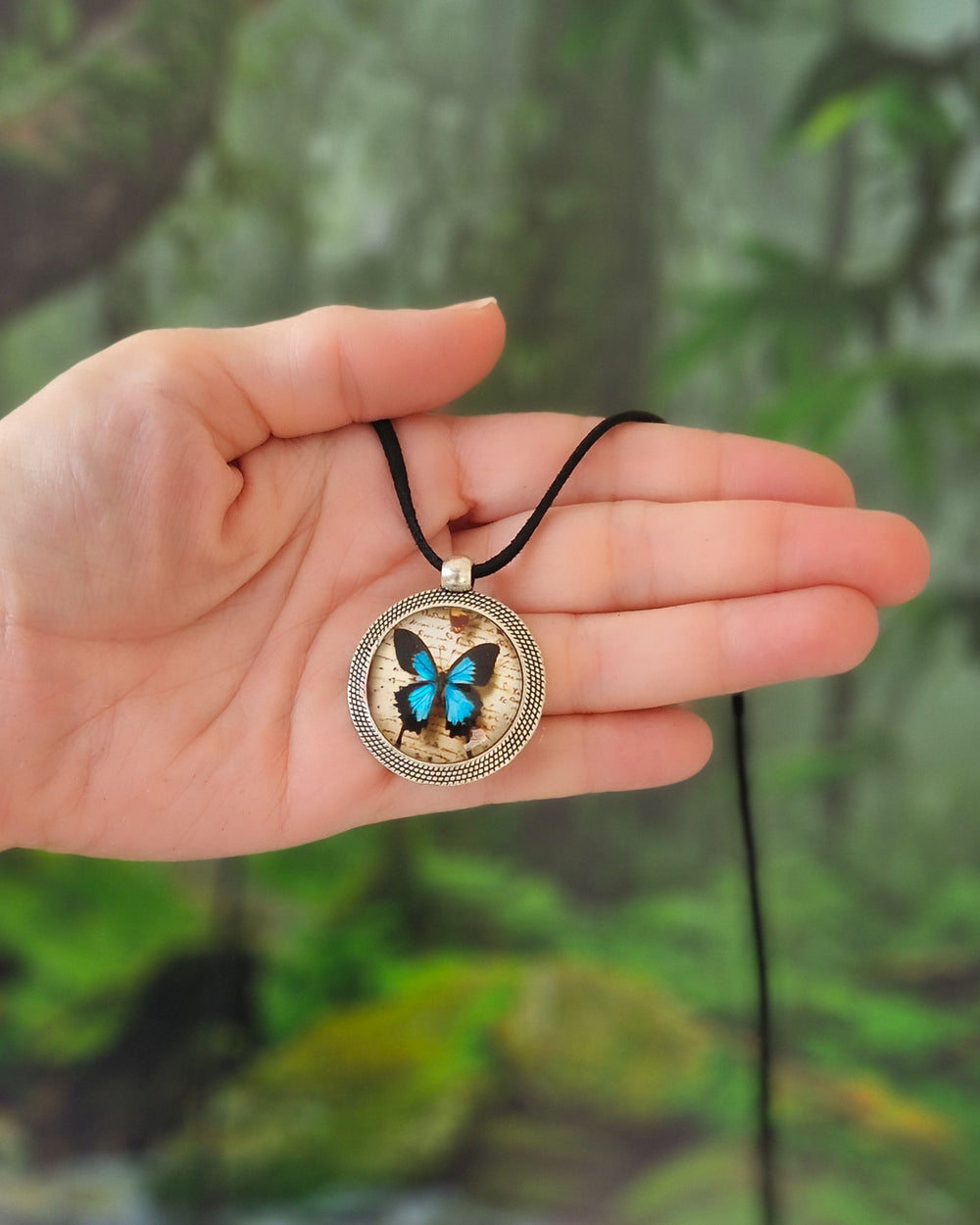 Hand holding Ulysses butterfly necklace with blue butterfly pendant, silver frame and black cord with a tropical rainforest background