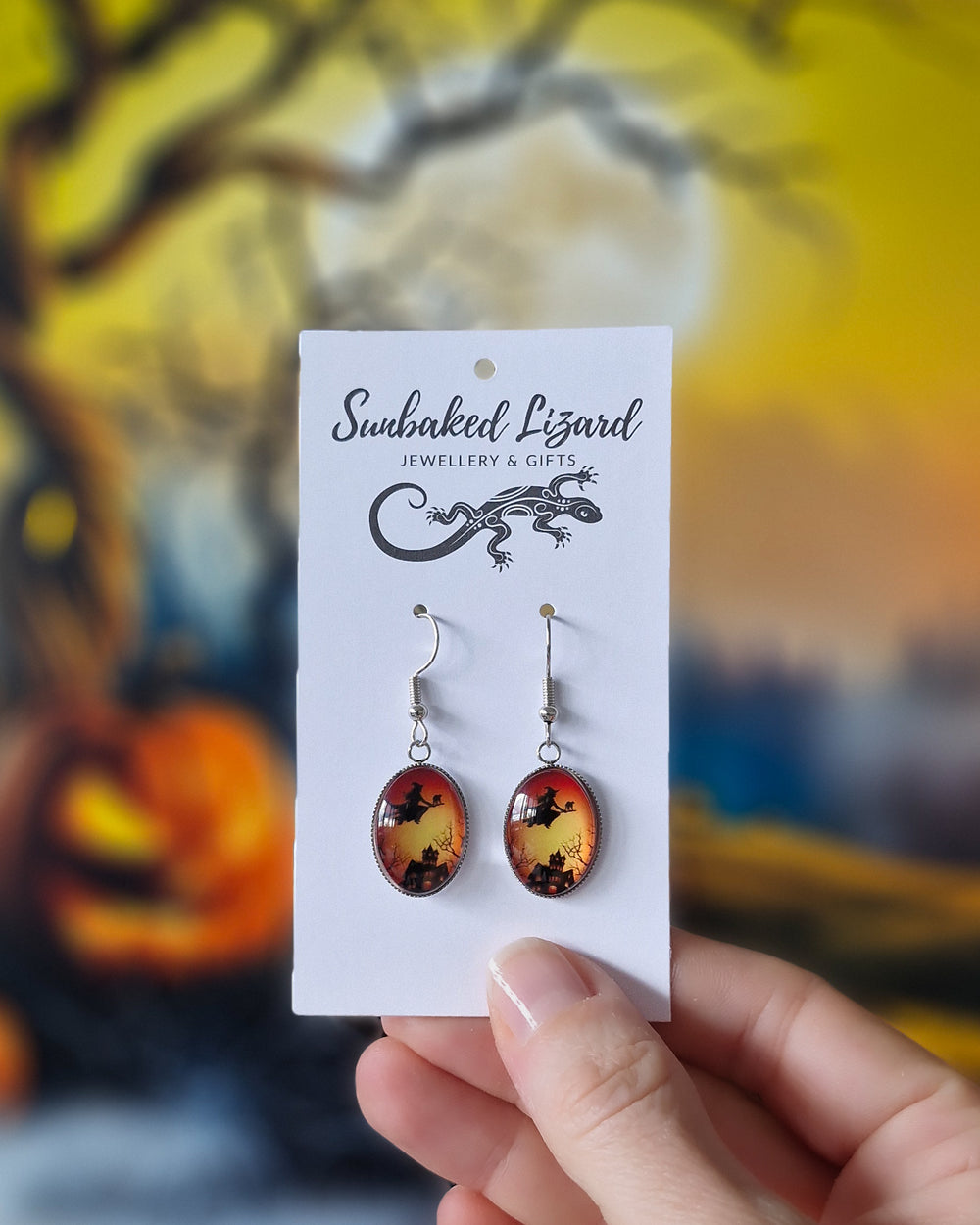 Hand holding Glass oval Halloween earrings showing witch and cat flying over haunted house at sunset, on a jewellery backing card to show scale