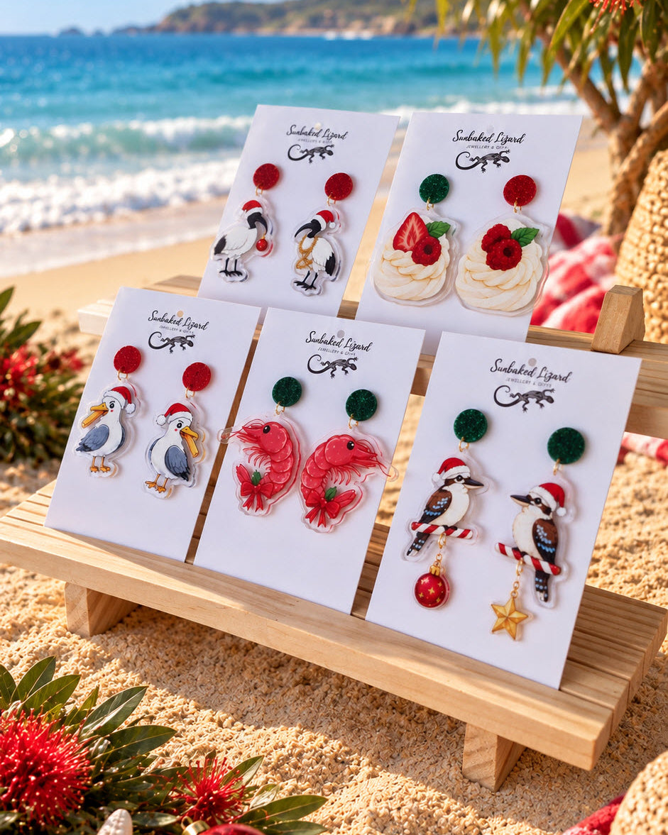 Five pairs of Australian Christmas earrings on Sunbaked Lizard cards at the beach, featuring an ibis, pavlova, seagull, prawn, and kookaburra