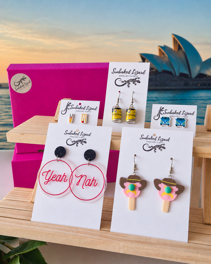 Australiana Earrings Gift Box, five pairs of Aussie earrings on Sunbaked Lizard cards, including yeah nah hoops, a retro van, and cricket stumps, with the Sydney Opera House at sunset behind
