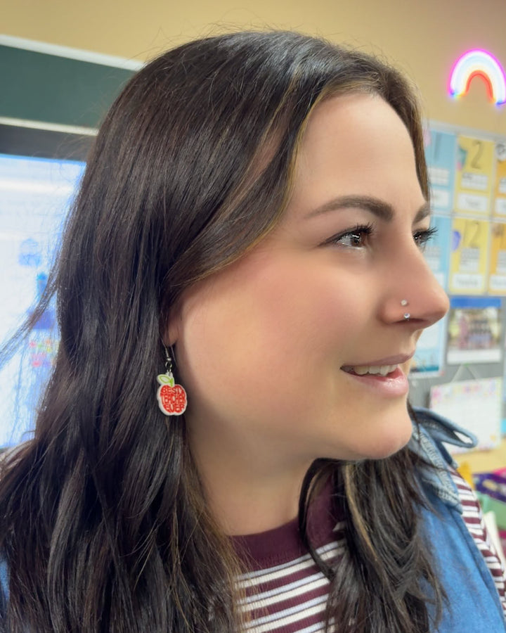 Side view of Best Teacher Ever Earrings featuring a red apple shape, green leaf and red lettering, worn in a colourful classroom.