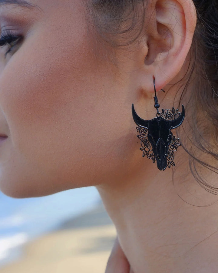 Model wearing black filigree bull skull drop earrings with floral detail at the beach