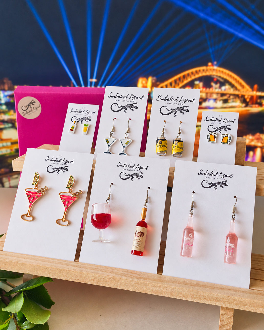 Booze Earrings Gift Box, seven pairs of cocktail, wine, beer, and spirit earrings on Sunbaked Lizard cards beside a pink gift box, with the Sydney Harbour Bridge lit up behind