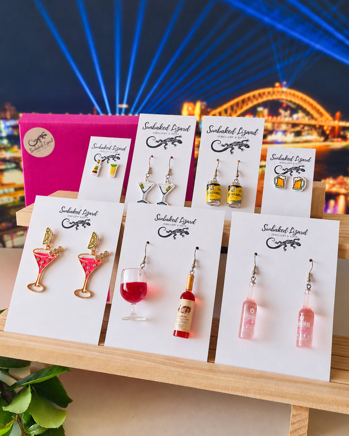Booze Earrings Gift Box, seven pairs of cocktail, wine, beer, and spirit earrings on Sunbaked Lizard cards beside a pink gift box, with the Sydney Harbour Bridge lit up behind