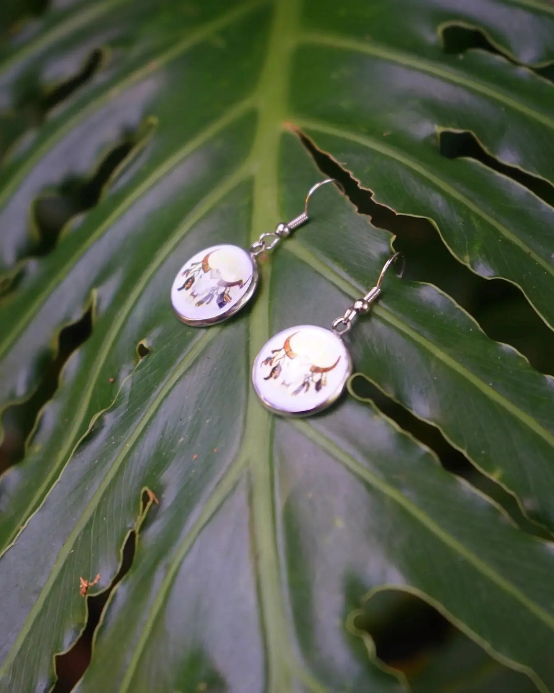 Small round bull skull drop earrings with silver hooks resting on a green leaf