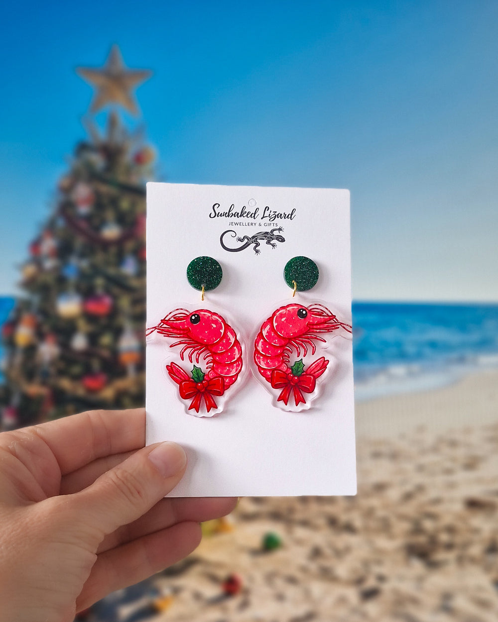 Christmas prawn earrings with green glitter tops, red prawns, bows and holly on a beach Christmas background.