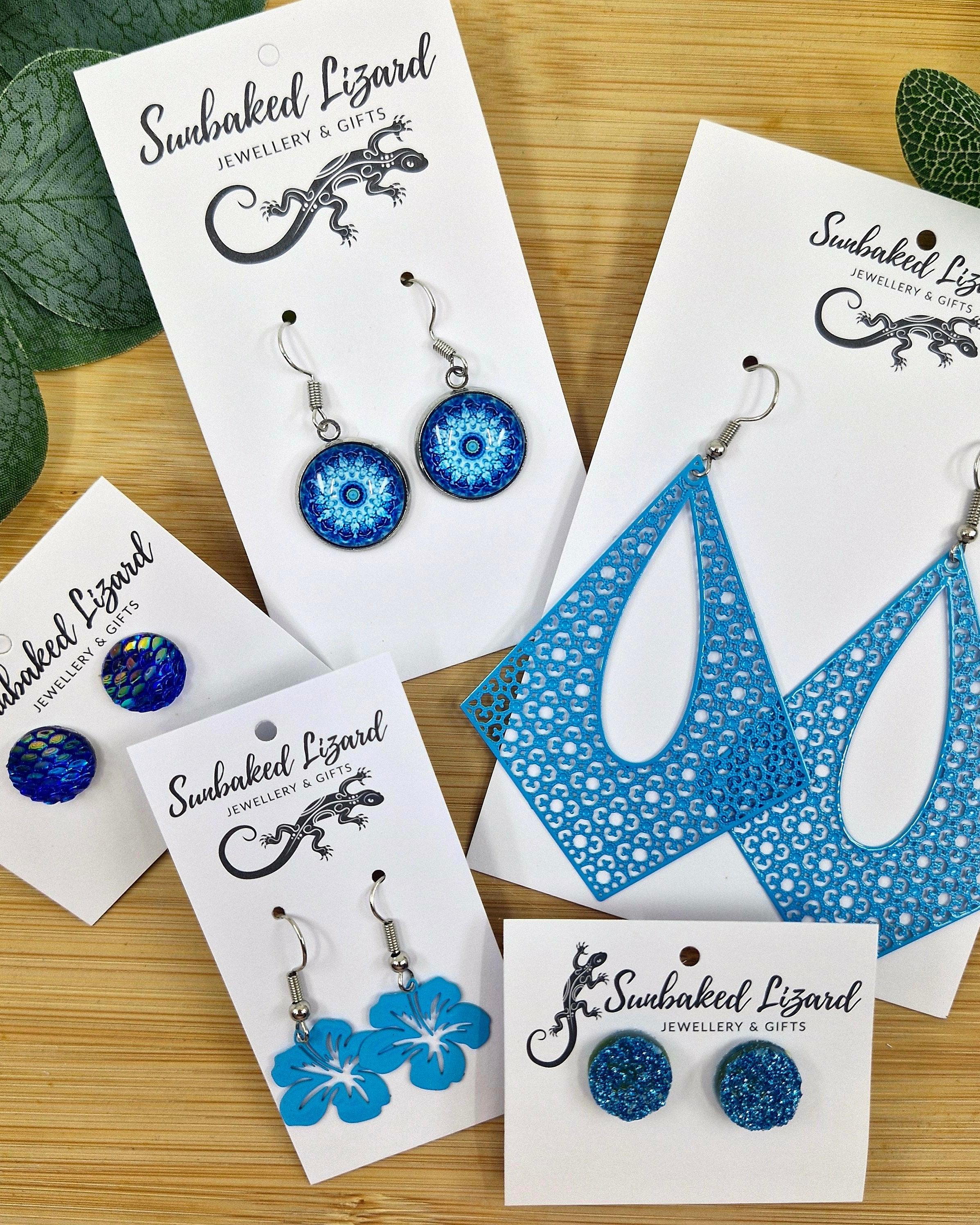 Selection of bright blue earrings from Sunbaked Lizard, including statement drop earrings, floral designs, and blue stud styles on a wooden background