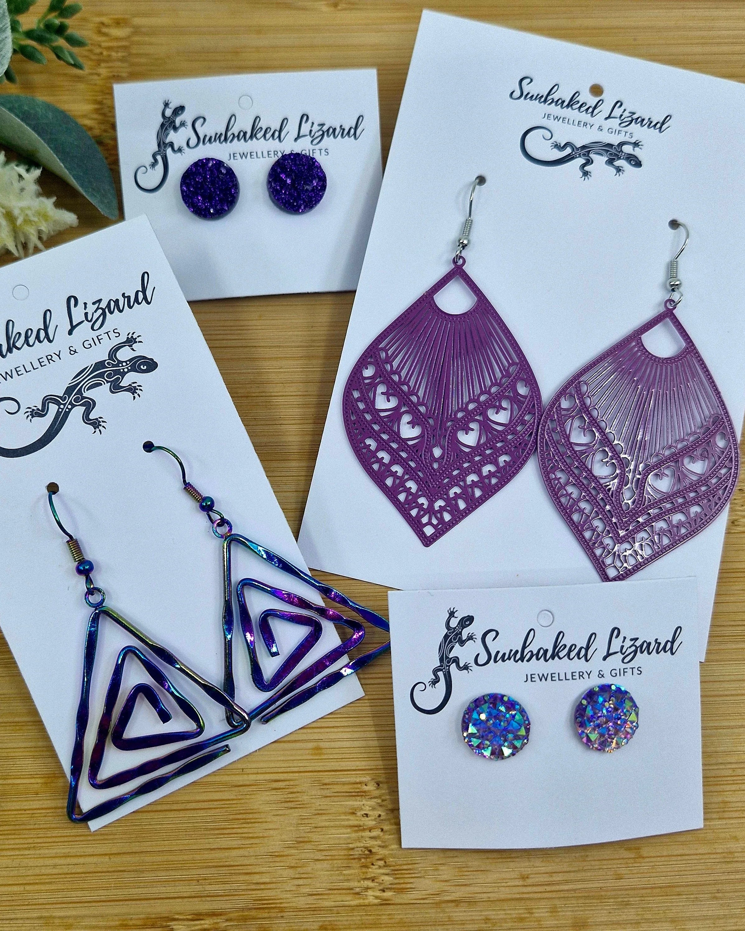 Selection of purple earrings from Sunbaked Lizard, featuring glitter stud earrings, filigree drop designs, and iridescent geometric styles on a wooden background