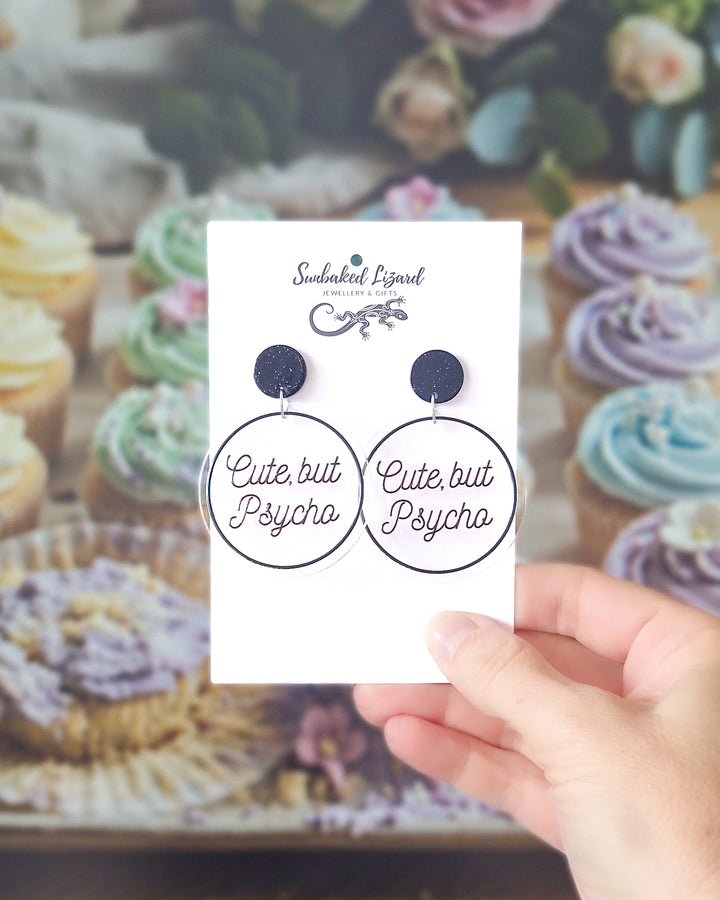Cute But Psycho black acrylic statement earrings displayed on a Sunbaked Lizard card with pastel cupcakes behind.