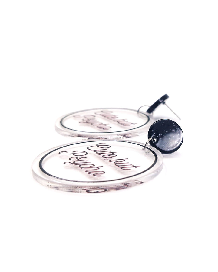 Close-up of Cute But Psycho acrylic statement earrings with black script, clear round charms and glitter black studs