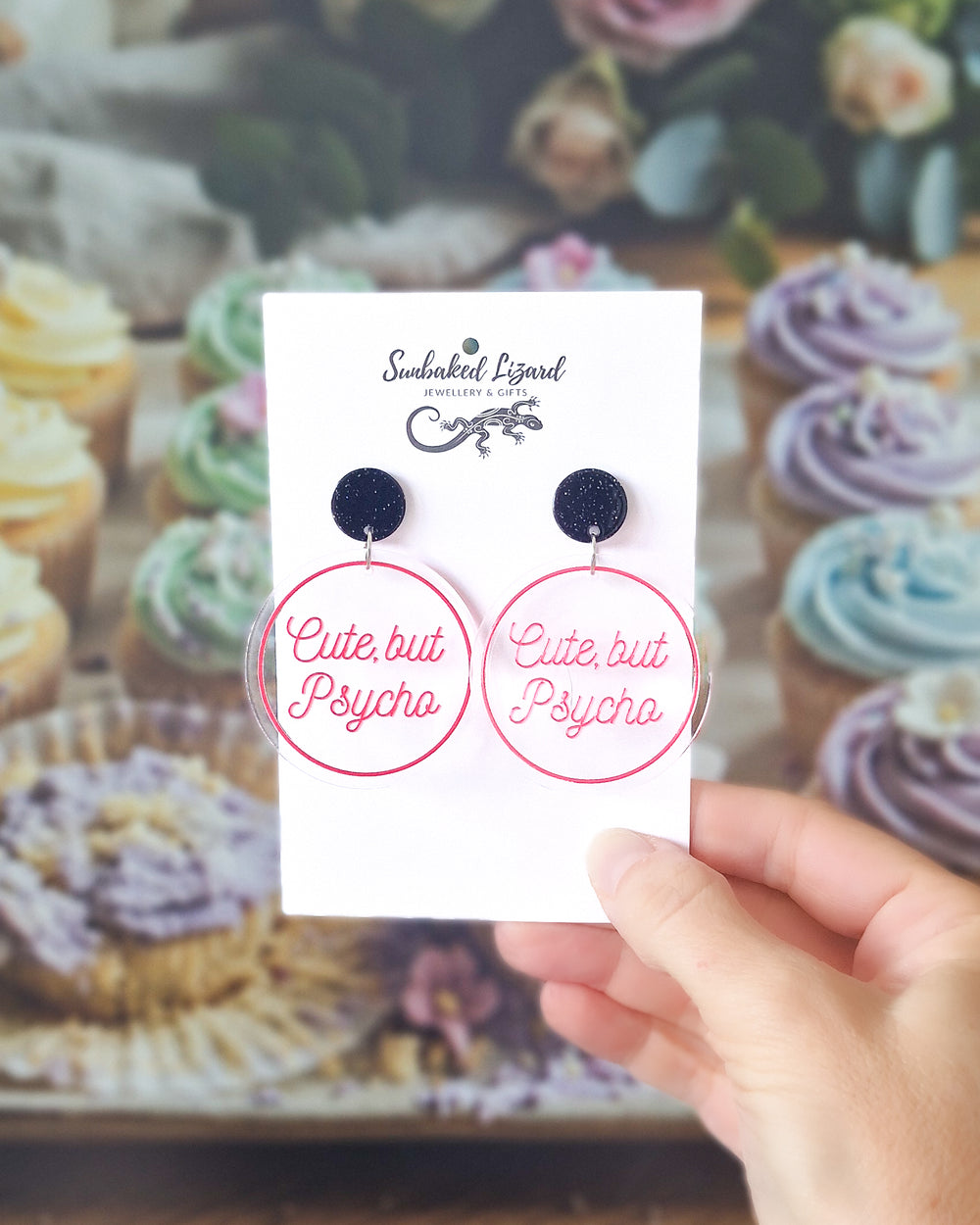 Cute But Psycho pink acrylic statement earrings displayed on a Sunbaked Lizard card with pastel cupcakes behind