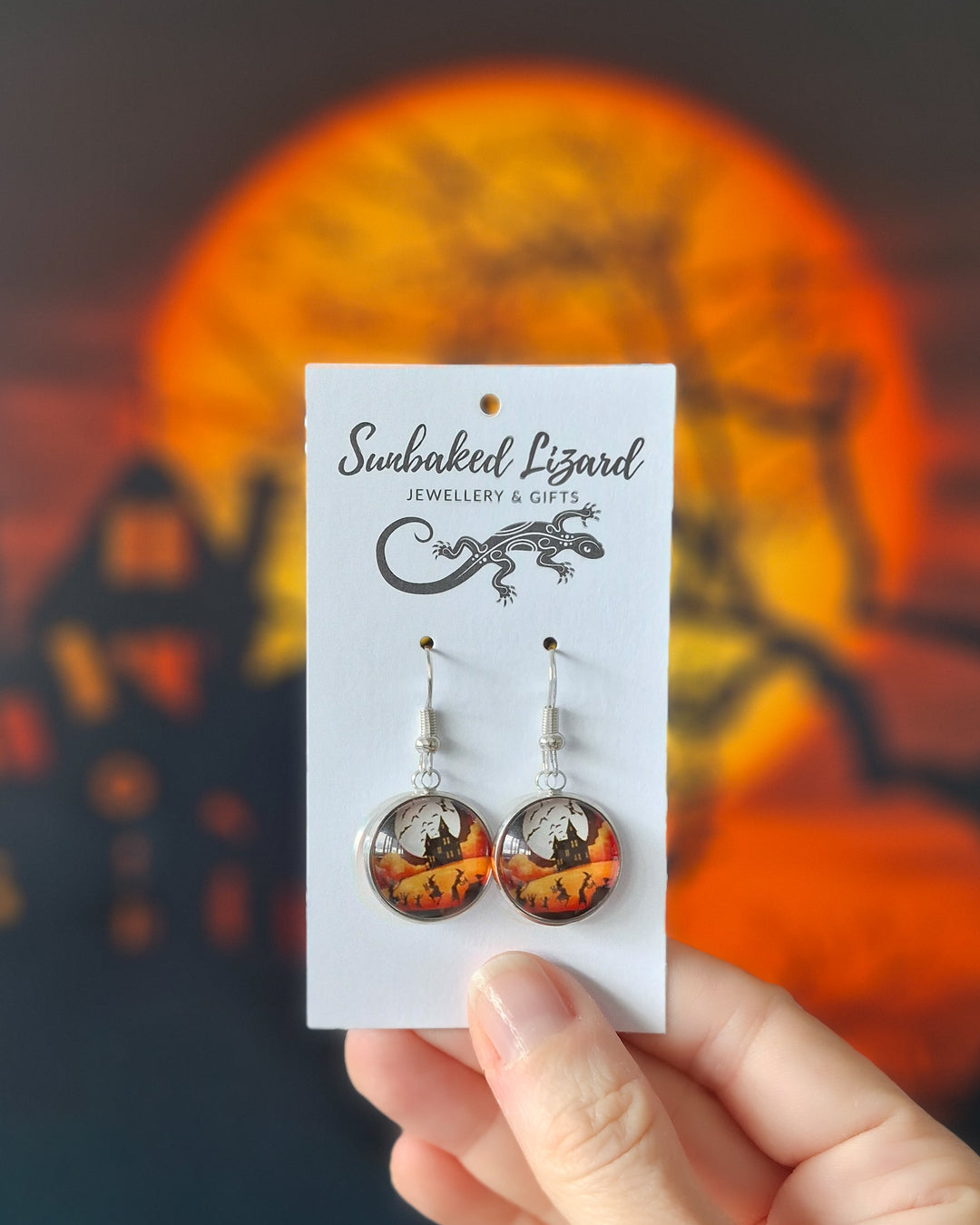 Round glass Halloween earrings on a Sunbaked Lizard jewellery card showing witches dancing before a haunted house under a full moon, silver hooks