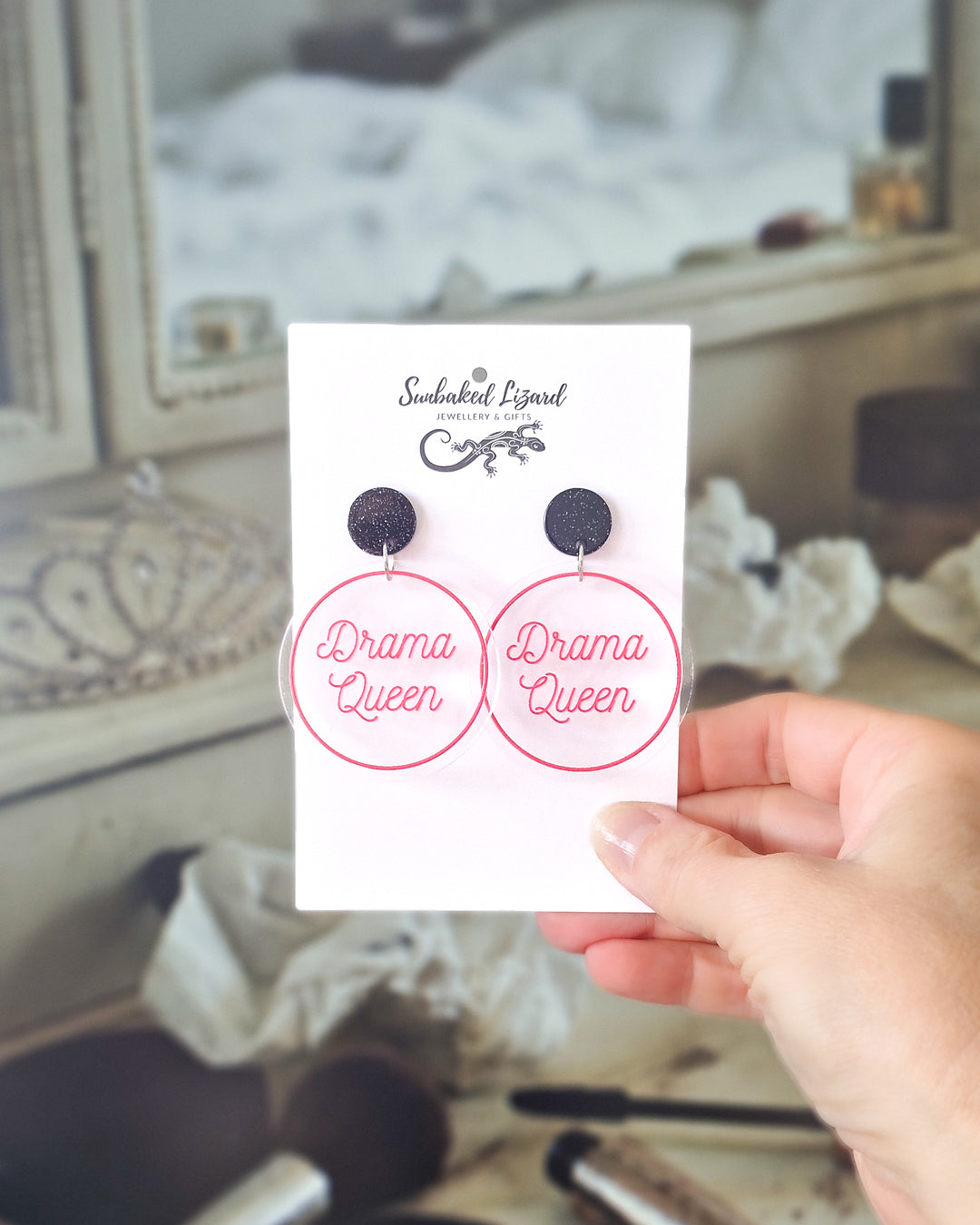 Drama Queen pink acrylic statement earrings displayed on a Sunbaked Lizard card at a vanity table