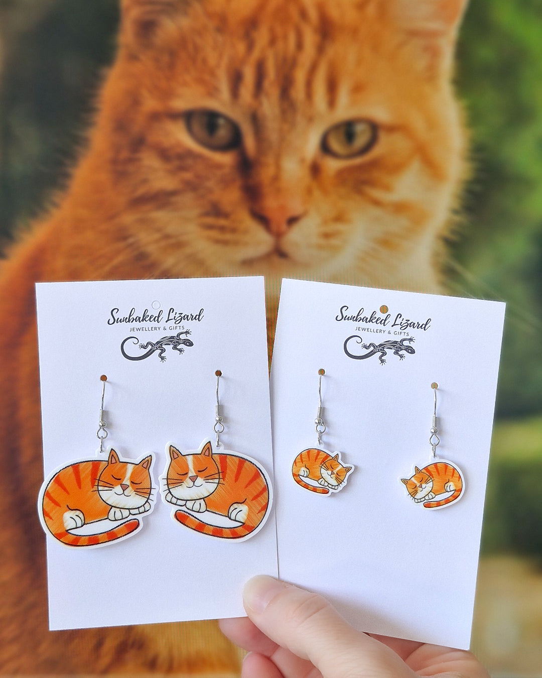 Ginger cat earrings in two sizes displayed on Sunbaked Lizard cards, held in front of a real ginger cat.