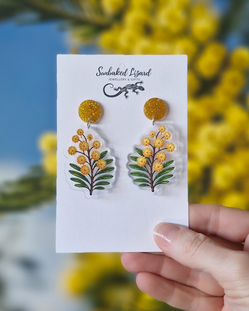 Golden wattle earrings on a Sunbaked Lizard card, held in front of a flowering wattle