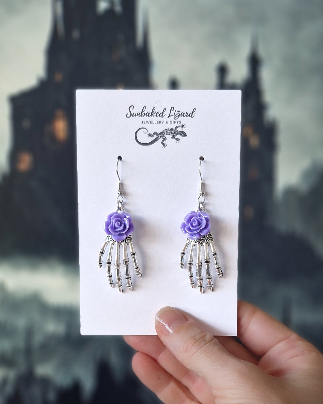 Halloween skeleton hand earrings displayed on a Sunbaked Lizard card showing scale with purple roses and a spooky gothic background