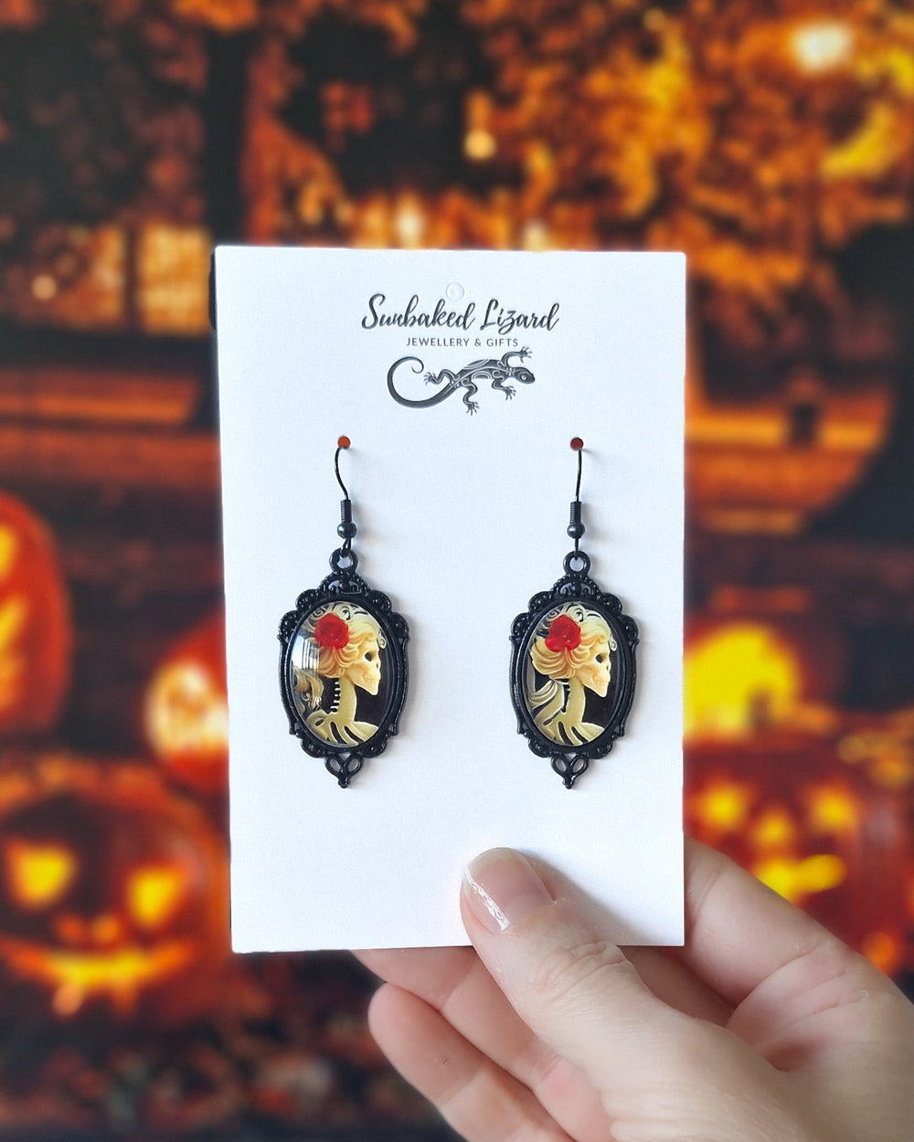 Gothic skeleton cameo Halloween earrings displayed on a Sunbaked Lizard card with a spooky pumpkin Halloween background