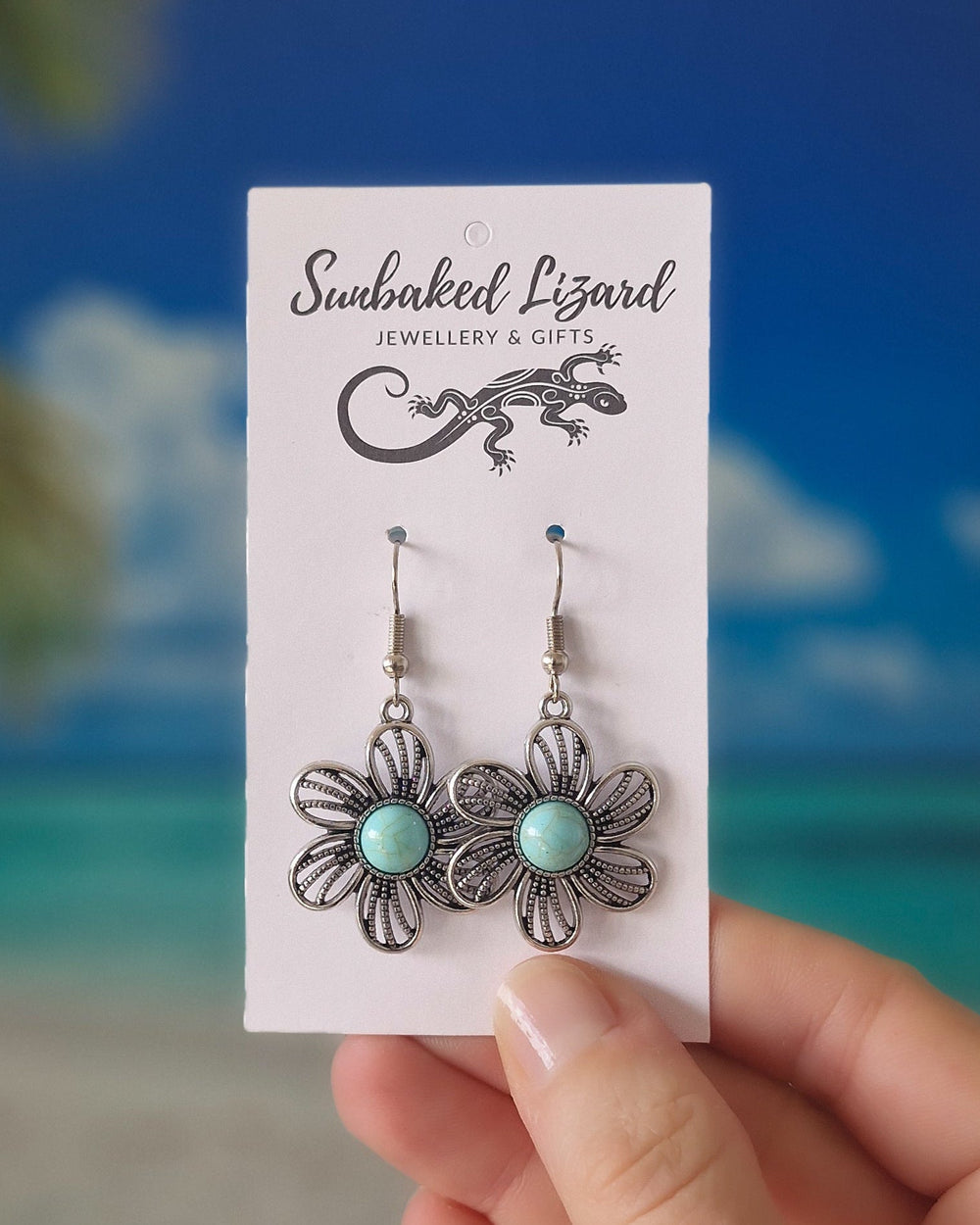 A pair of silver filigree flower drop earrings with light blue-green turquoise centers, displayed on a white 'Sunbaked Lizard Jewellery & Gifts' card, held by a hand against a blurred beach background.