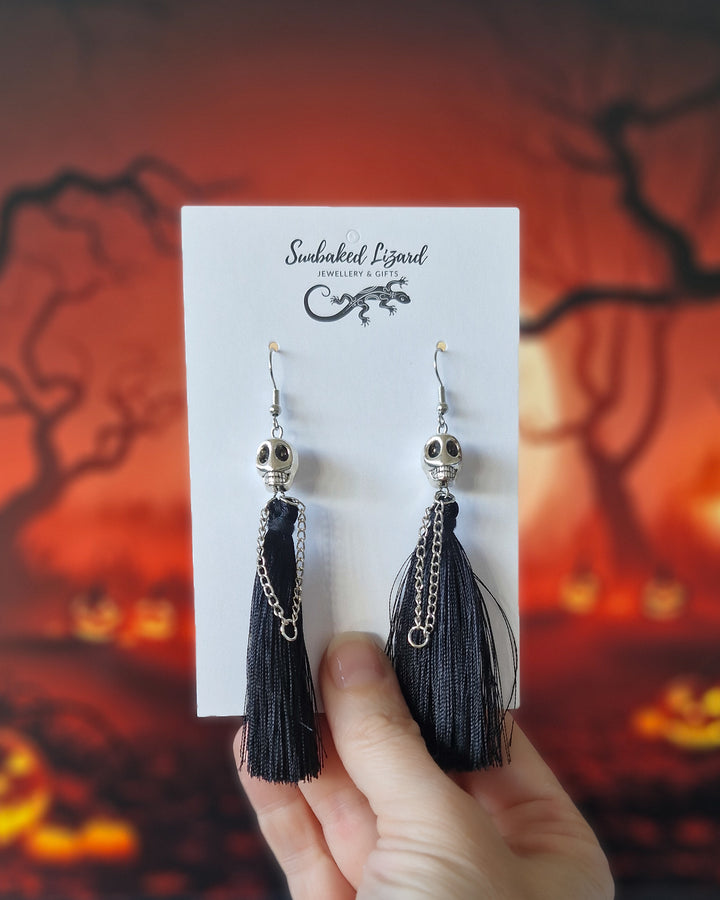 Halloween skull tassel earrings displayed on a Sunbaked Lizard card with long black tassels and a spooky pumpkin background