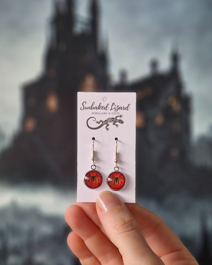 Halloween spider drop earrings with glossy red cabochon pendants, black spider silhouettes held on a jewellery backing card with a spooky background showing scale