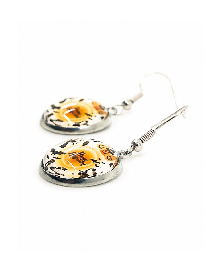 Close up showing detail of round glass Halloween earrings with pumpkin characters, bats and Happy Halloween text on white background, silver hooks