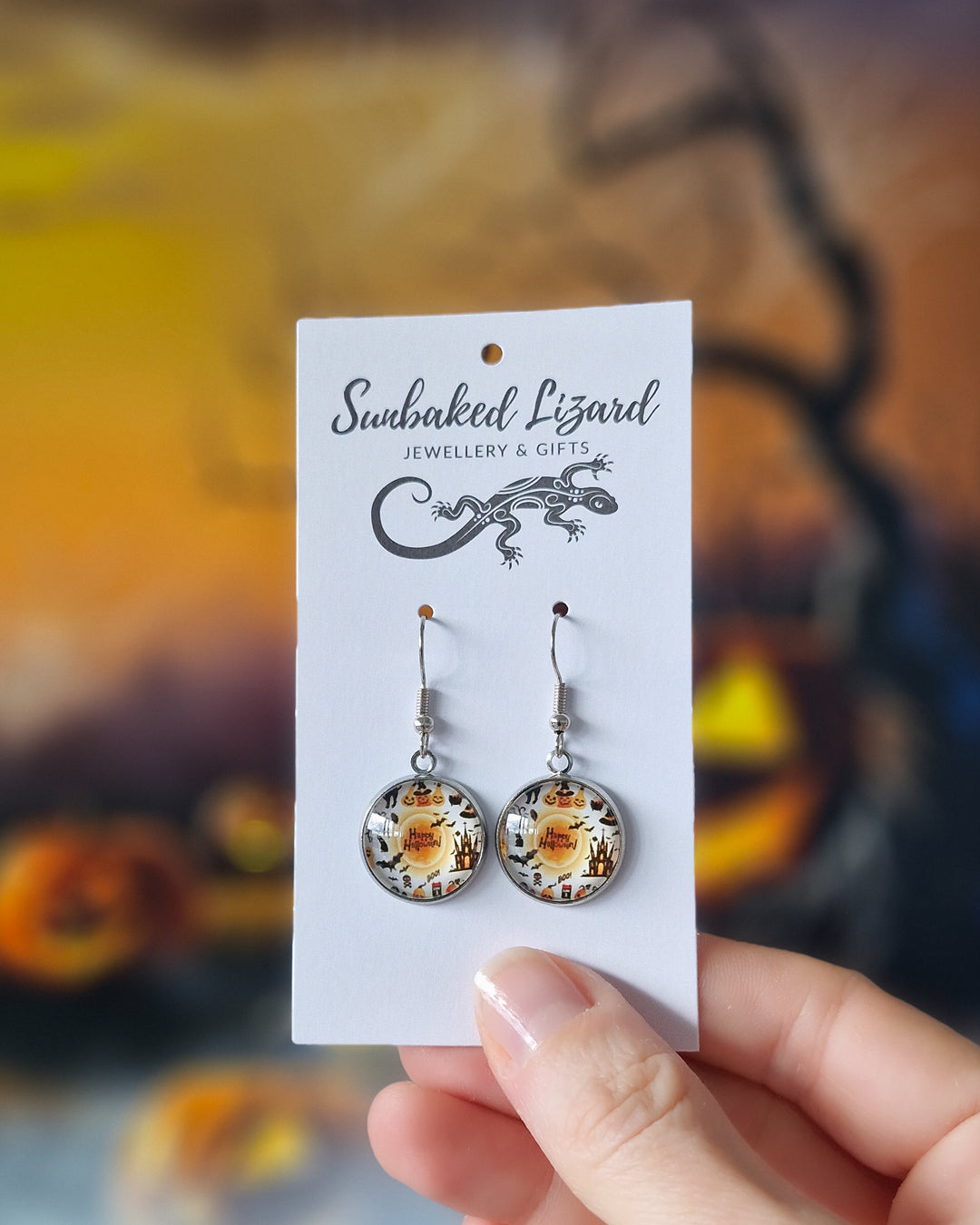 Round glass Halloween earrings with pumpkin characters, bats and Happy Halloween text on a Sunbaked Lizard jewellery card with Halloween pumpkins in the background