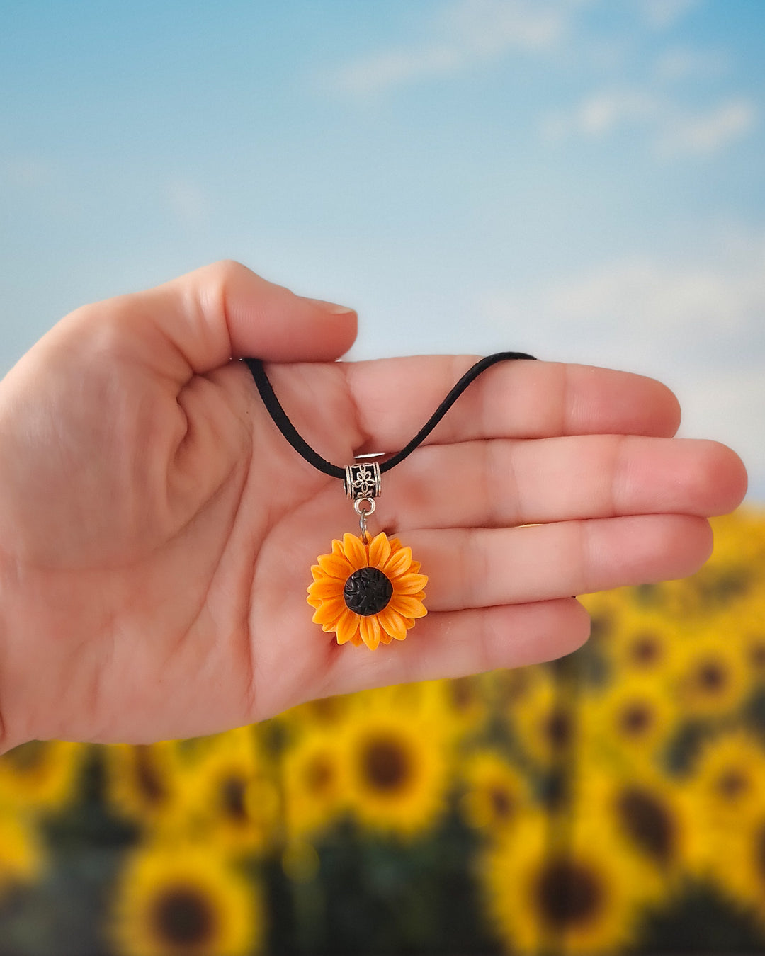 Hand holding a sunflower pendant necklace with a blurred sunflower field in the background
