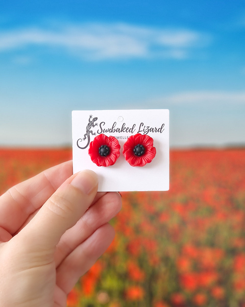 Hand holding red poppy flower stud earrings on a jewellery display card with a soft-focus poppy field in the background.