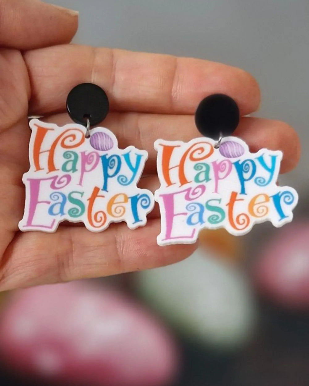 Pair of 'Happy Easter' earrings held between fingers with blurred Easter eggs in the background.
