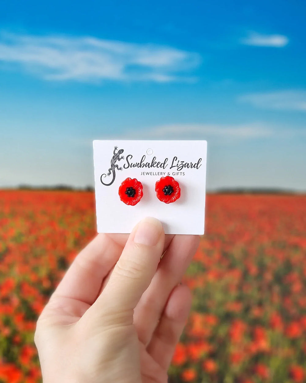 Red poppy flower stud earrings on a Sunbaked Lizard jewellery card, held in front of a blurred poppy field and blue sky.