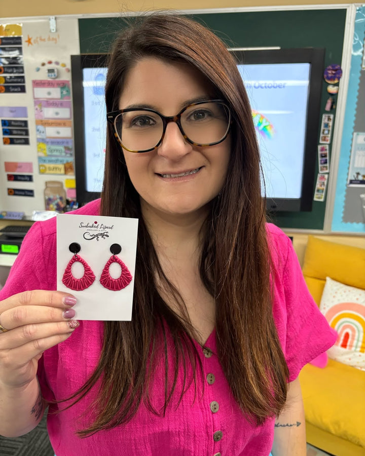 Pink Acrylic Woven Earrings with black tops displayed on a Sunbaked Lizard product card in a classroom.