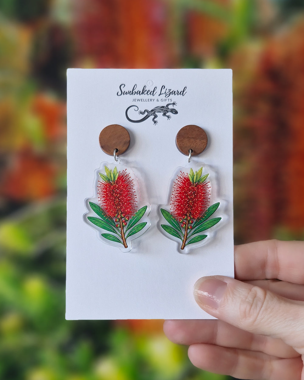 Red bottlebrush earrings on a Sunbaked Lizard card, held in front of a flowering bottlebrush