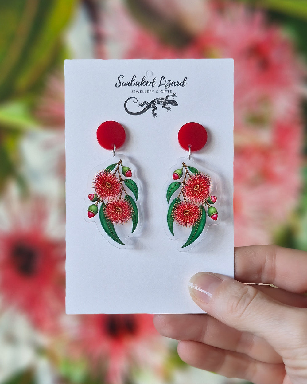 Red gum blossom earrings on a Sunbaked Lizard card, held in front of a flowering gum