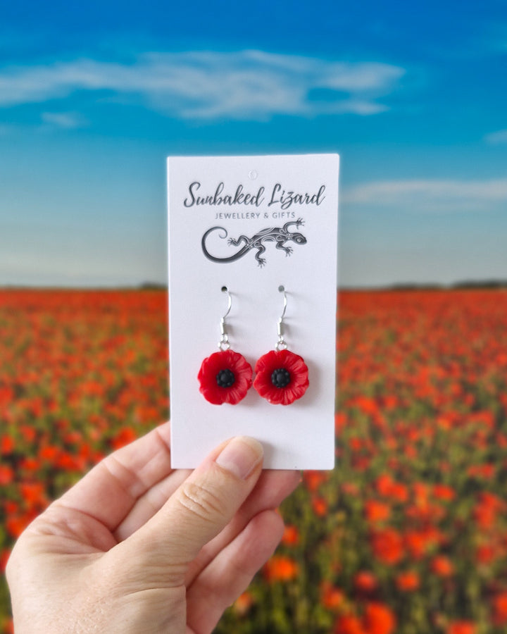 Red poppy earrings shown in stud and dangle styles on Sunbaked Lizard display cards against a blurred poppy field background.