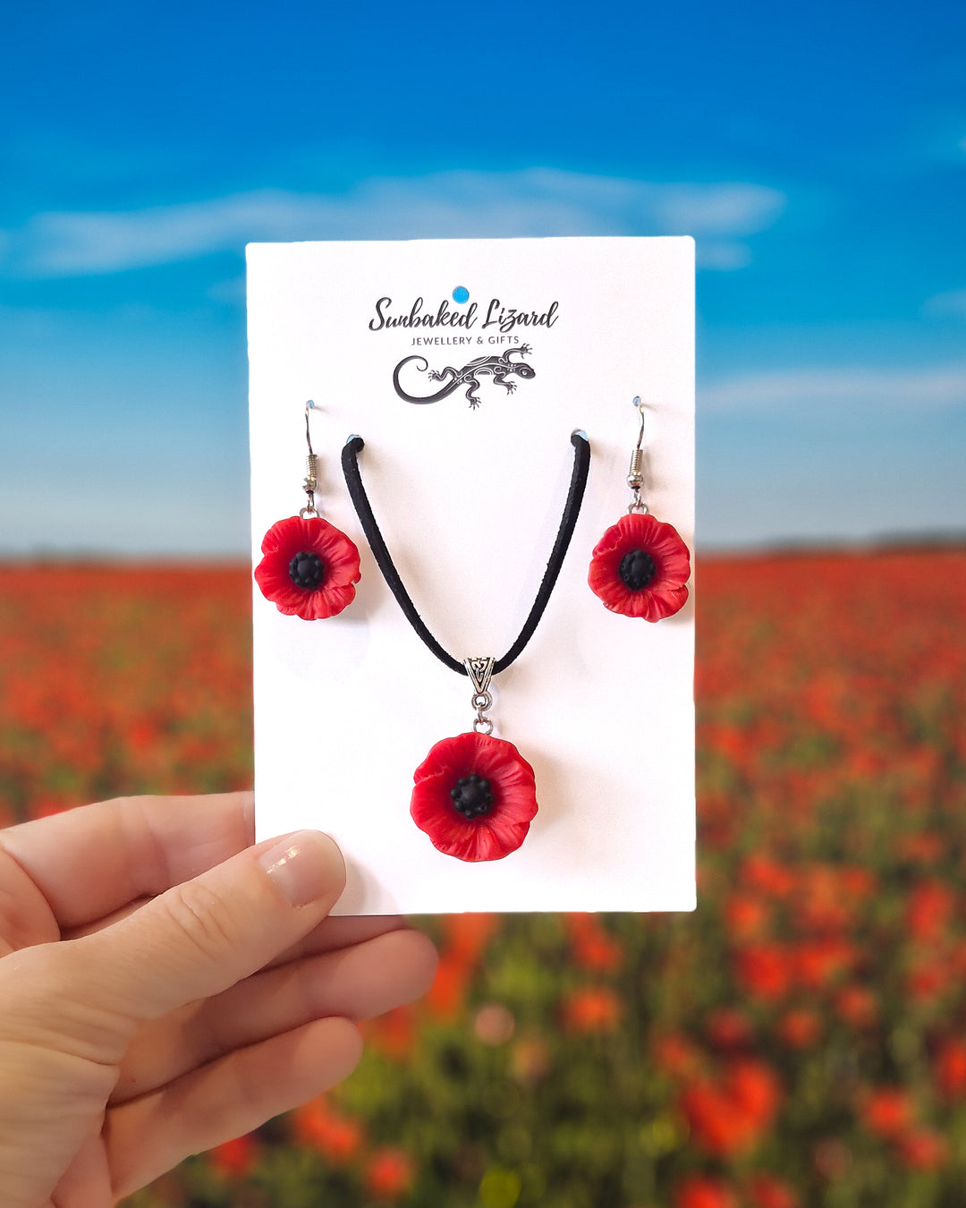 Red poppy necklace and matching drop earrings set on Sunbaked Lizard card with poppy field background