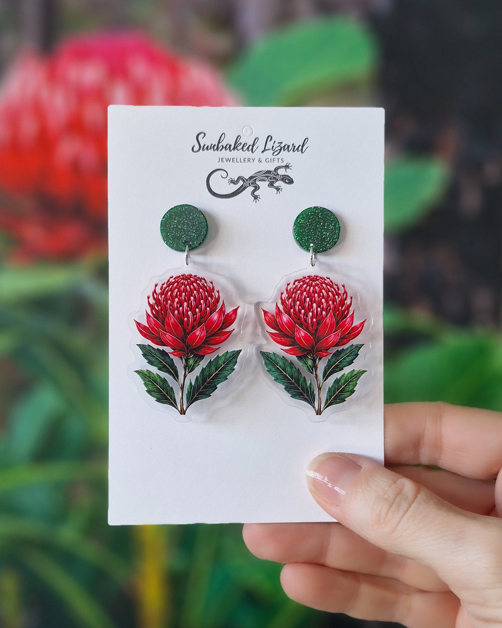 Red waratah earrings on a Sunbaked Lizard card, held in front of a flowering waratah