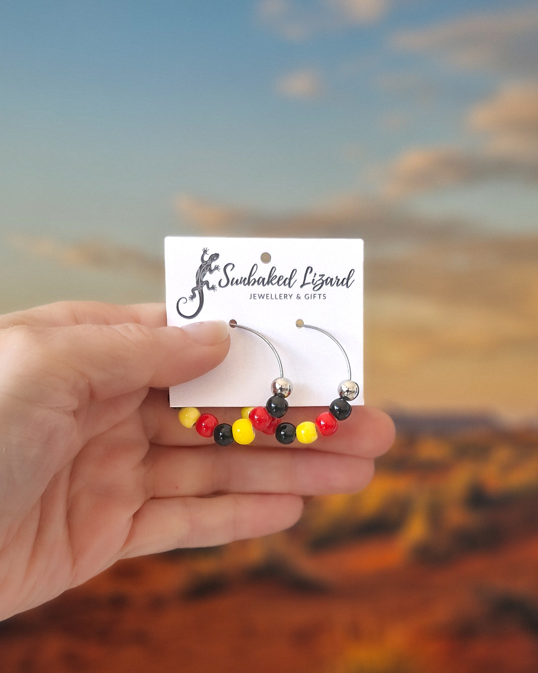 Hand holding a pair of red, yellow and black Indigenous inspired hoop earrings with glossy beads on silver hoops on a card with a blurred desert landscape in the background.