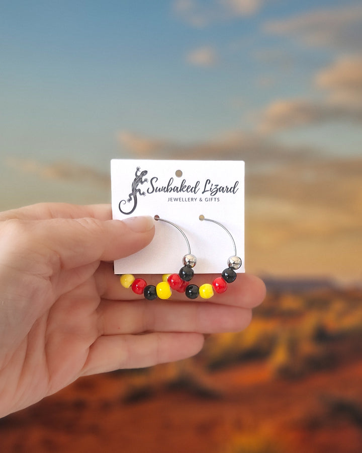 Hand holding a pair of red, yellow and black Indigenous inspired hoop earrings with glossy beads on silver hoops on a card with a blurred desert landscape in the background.