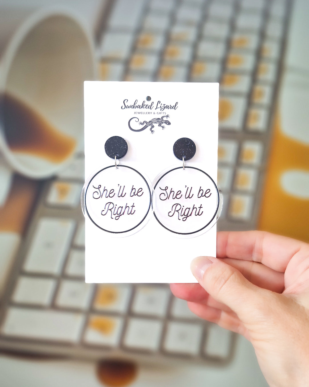 She’ll Be Right black acrylic statement earrings displayed on a Sunbaked Lizard card with coffee spilled into a keyboard behind.