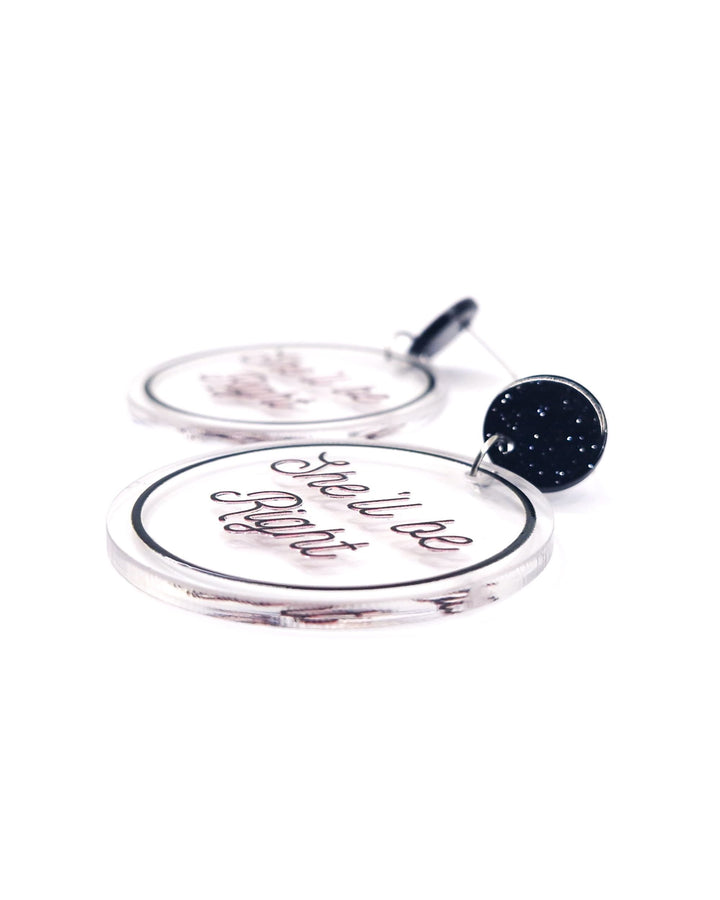 Close-up of She’ll Be Right acrylic statement earrings with black script, clear round charms and glitter black studs.