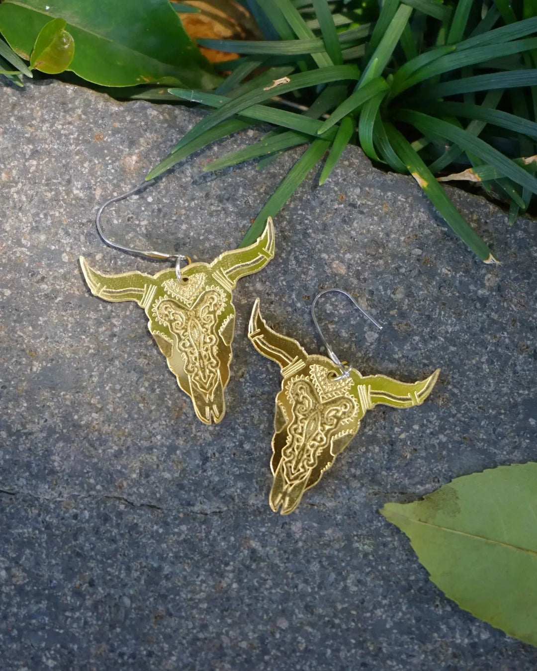 Shimmer gold bull head dangle earrings with engraved western detail displayed on stone with greenery.