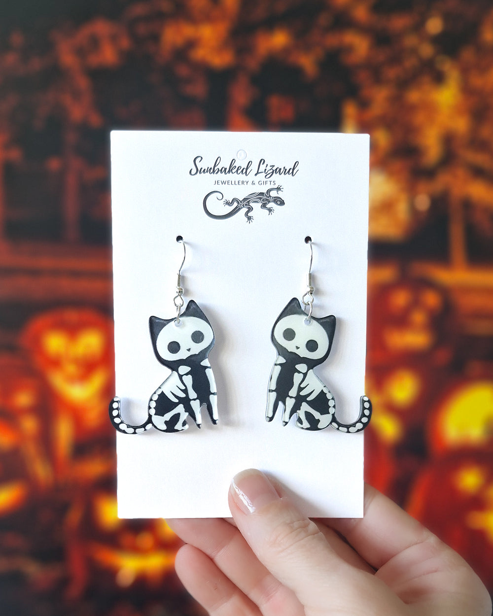 Skeleton cat Halloween earrings displayed on a Sunbaked Lizard card showing scale with a spooky pumpkin Halloween background