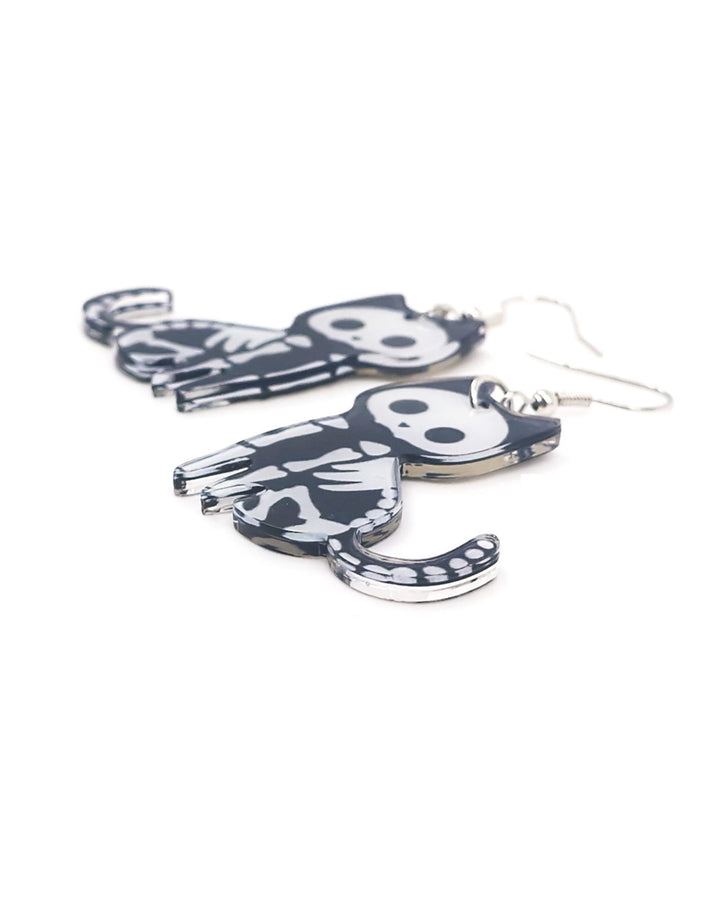 Close up photo of  Black and white skeleton cat Halloween earrings with curled tails and silver hooks, laid flat on a white background